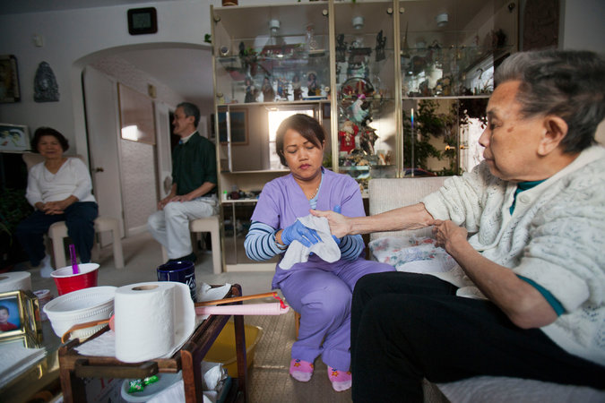 Savan Mok, a home health aide, assisting Oun Oy, 90, right, who had a stroke in 2012. Ms. Oy is from Cambodia and lives in Jenkintown, Pa., with her son and his wife, at rear. Jessica Kourkounis for The New York Times