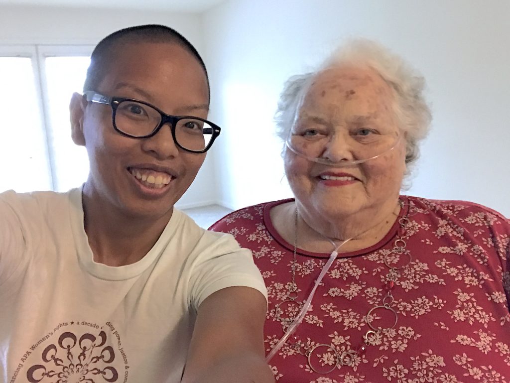 liz and Mom take a selfie in the living room of their new apartment together this past September. liz is a dark-skinned, Vietnamese person with black shaved head and black glasses. They wear a light blue NAPAWF t-shirt. Mom is an older, light-skinned Caucasian with short, white, wavy hair. She wears a red short-sleeved cotton shirt with small, white flowers. She wears an oxygen cannula.