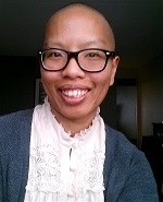 A torso shot of a dark-skinned, Vietnamese person with shaved black hair and black glasses smile. liz wears a ruffled, dusty pink dress with a heather gray, long-sleeved sweater.