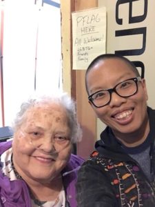 Liz, a dark-skinned, person with shaved black hair and black glasses, with Mom, a light-skinned white woman with short wavy white hair, smile in front of the PFLAG meeting room. A piece of notebook paper hangs outside the door, "PFLAG here; All welcome! LGBTQ+ friends family."